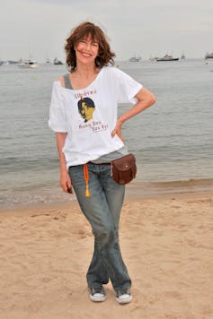 Një grua me një T-shirt, xhinse dhe një çantë në bel pozon në një plazh.