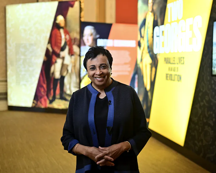 A middle-aged woman with light brown skin and dark hair stands and smiles with her hands clasped together.