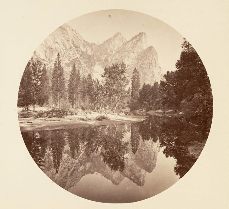 A sepia-toned image of a lake with massive trees and even bigger mountains behind it.