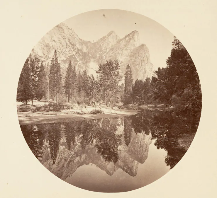 A sepia-toned image of a lake with massive trees and even bigger mountains behind it.
