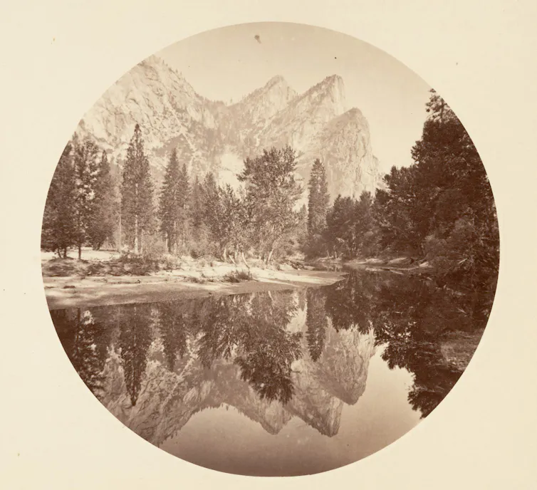 A sepia-toned image of a lake with massive trees and even bigger mountains behind it.