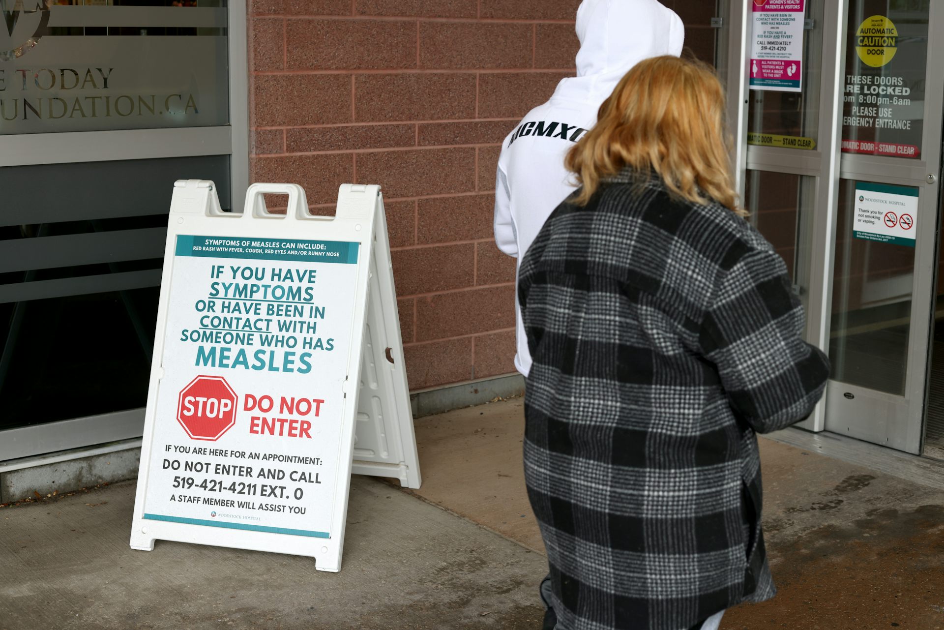 A sign reading 'If you have symptoms or have been in contact with someone who has measles STOP do not enter' outside a building entrace, with two people seen from behind