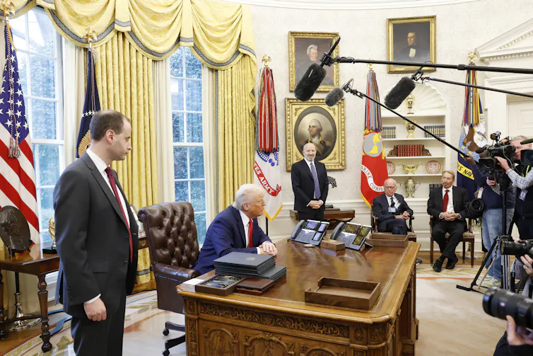 A wide shot of the Oval Office with Donald Trump at his desk and Rupert Murdoch in the corner