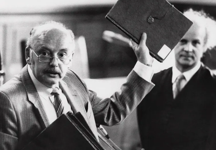 A man with glasses and a moustache holds up a black folder.