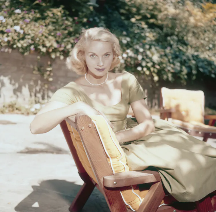 Saint sings on sun lounger in a garden, wearing a light green dress and a pearl necklace.