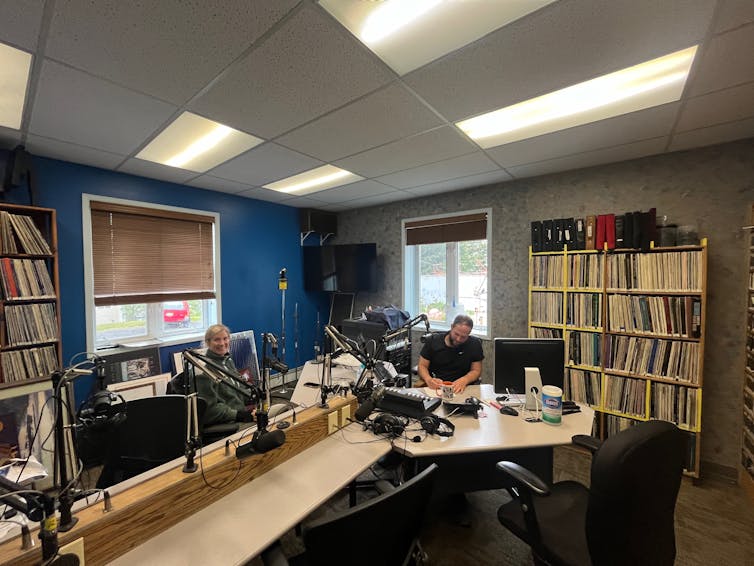 Dy persona brenda një studio radio, duke u ulur në një tryezë të gjatë-desk kombinim.
