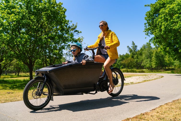 A woman riding a bike with a large cargo hold on the front which a child is sitting in.