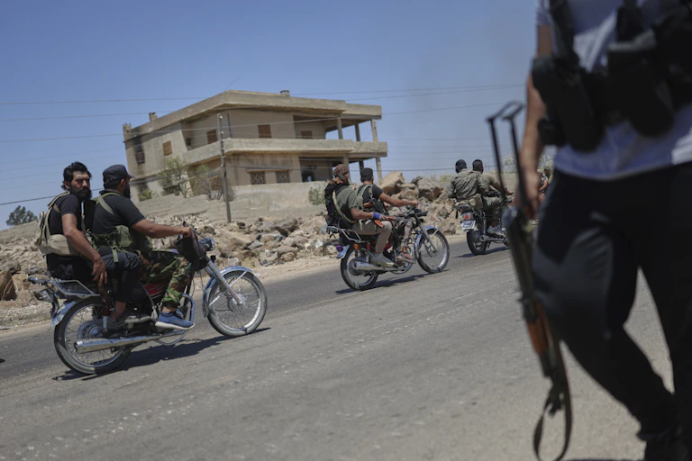 Armed men ride motorbikes on a street, while a man stands with a gun on the other side