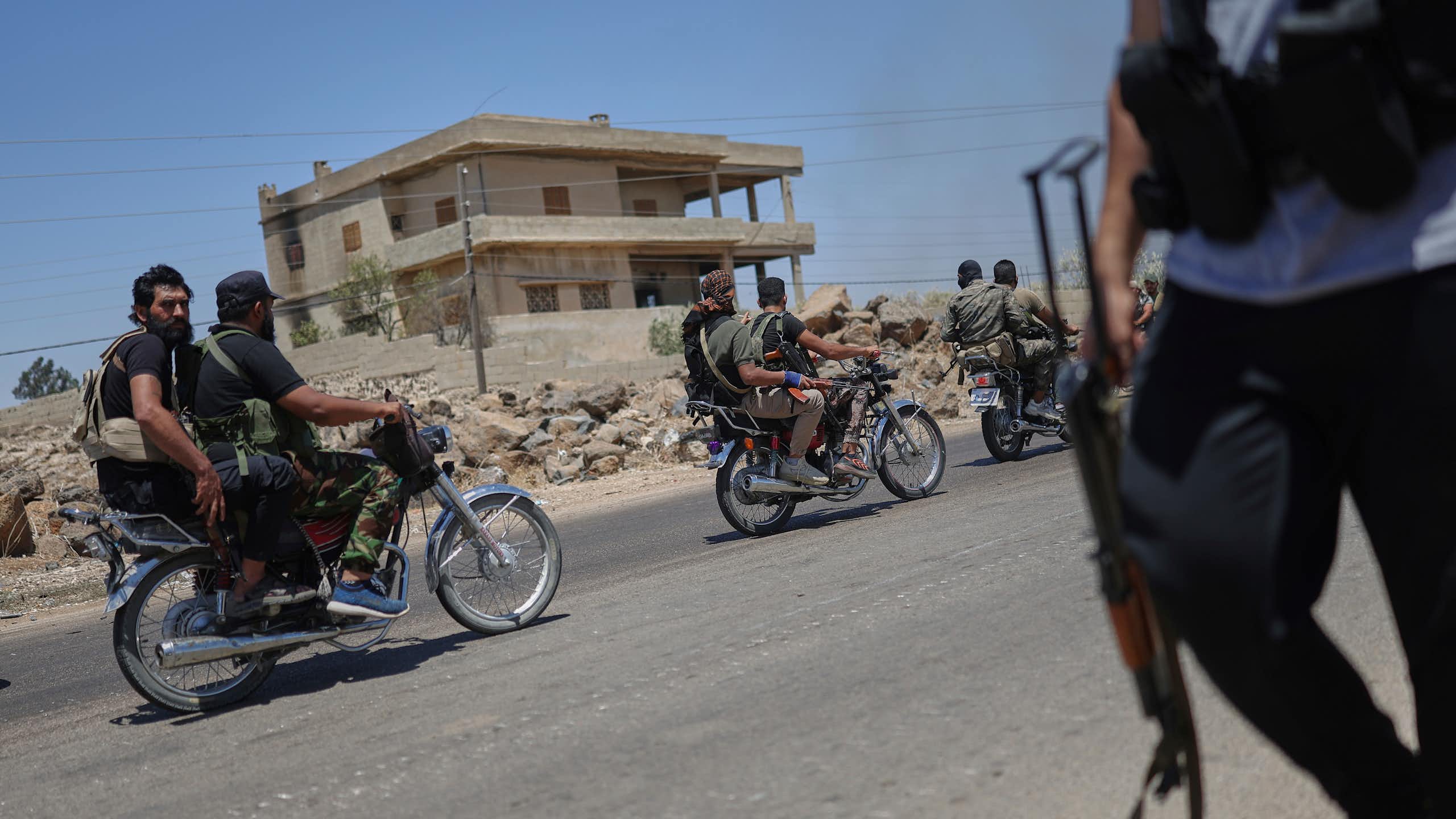 Armed men ride motorbikes on a street, while a man stands with a gun on the other side