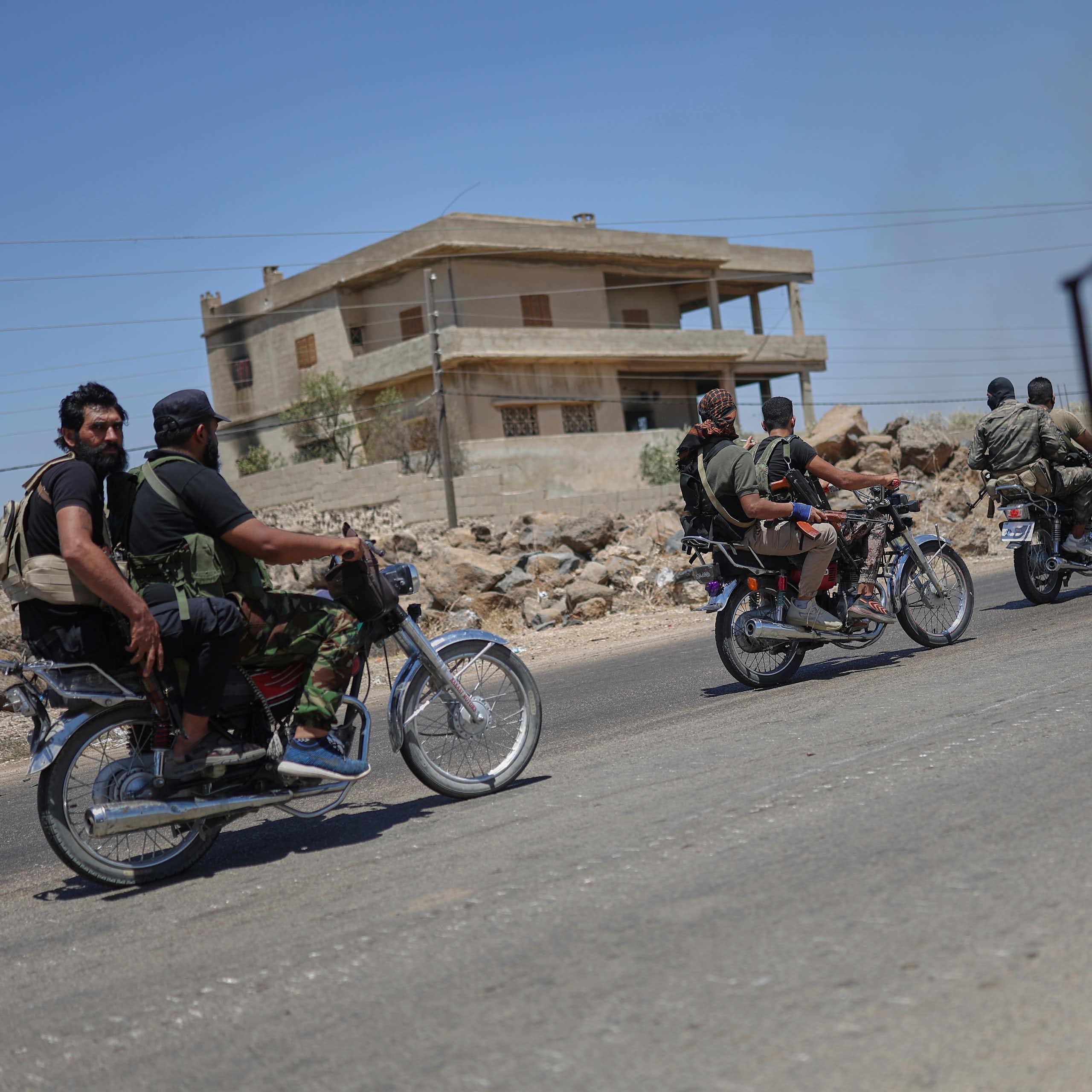 Armed men ride motorbikes on a street, while a man stands with a gun on the other side