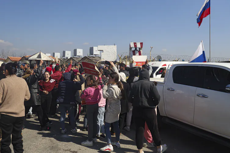 Several people, including women and children, stand next to parked vehicles.
