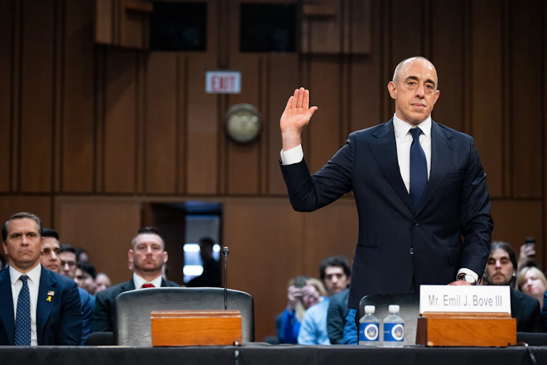 A bald, middle aged man with a navy blue suit, white shirt and navy tie stands in front of a placard that says Mr. Emil J. Bove III and raises his hand. People sit behind him in a room.