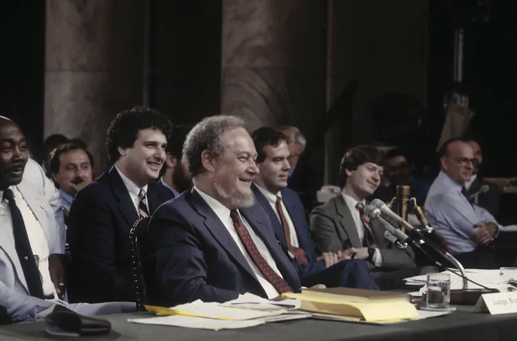 A man smiles and looks toward a microphone with people sitting behind him. All of them are dressed formally.