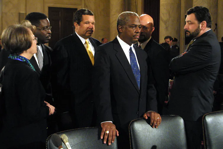 A man with brown skin and a black suit places a hand on a leather chair and stands alongside people dressed formally.