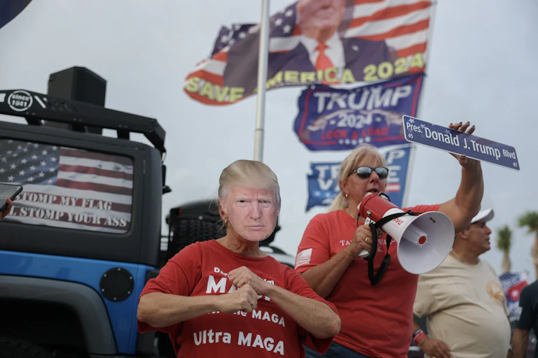 People wear red shirts and hold a sign that says 'Donald J. Trump' and a placard that looks like Trump's face. Flags that say Trump and show the president wave behind them.
