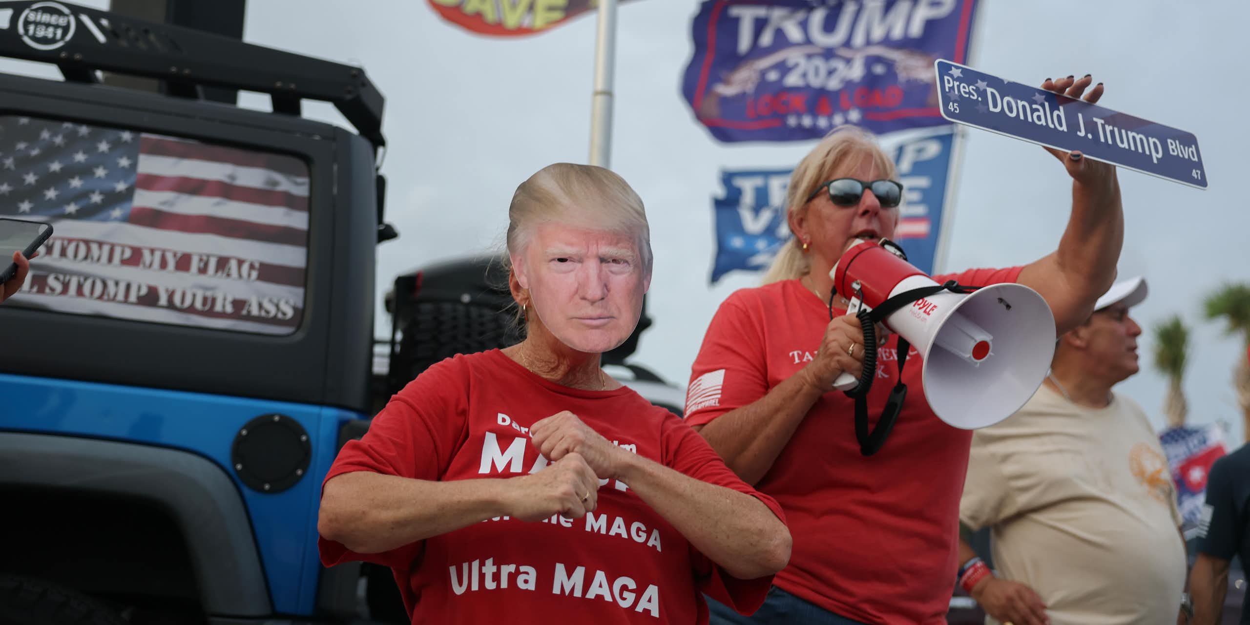 People wear red shirts and hold a sign that says 'Donald J. Trump' and a placard that looks like Trump's face. Flags that say Trump and show the president wave behind them.