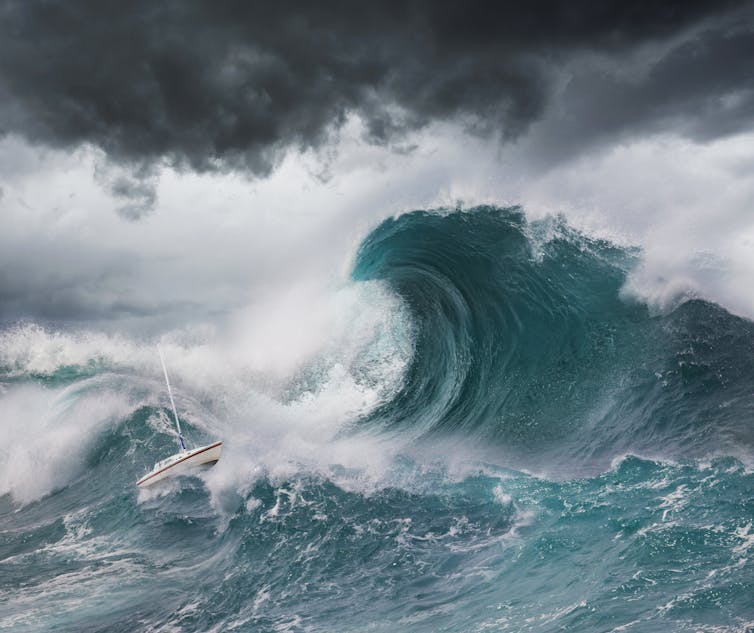 A sailboat caught in the swell of a tall wave, under a cloudy sky