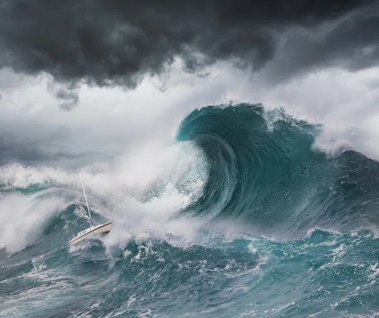 A sailboat caught in the swell of a tall wave, under a cloudy sky