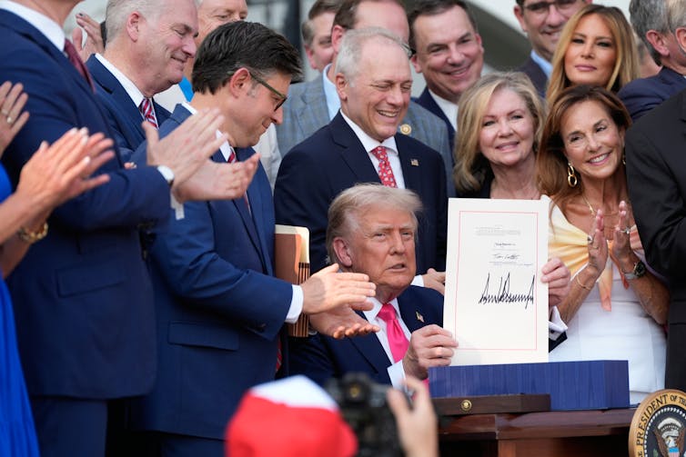 An older man with white hair sits and holds up a larger piece of paper. He is surrounded by people dressed formally who applaud and smile.
