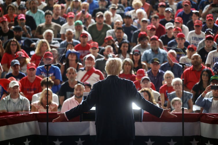 A large crowd of people sit and face board a man who is illuminated from the front.