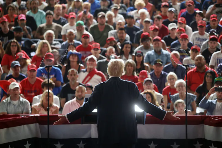 A large crowd of people sit and face board a man who is illuminated from the front.