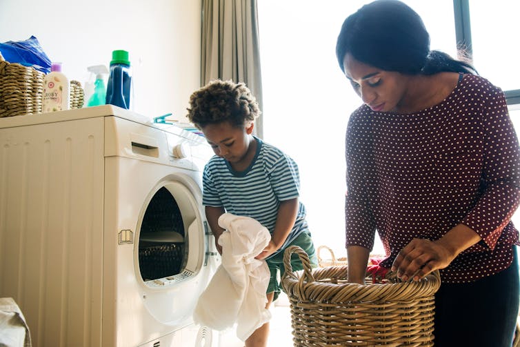 A woman puts laundry in a laundry basket. Beside her, a little boy takes some clothing out a dryer