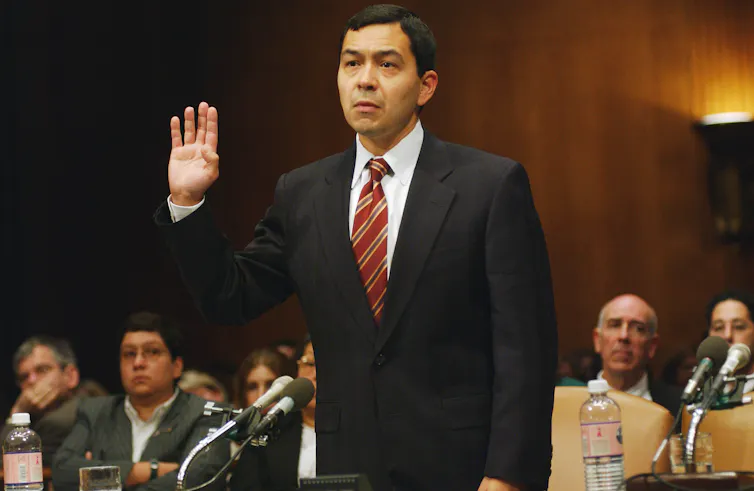 A dark-haired man in a suit, standing while swearing an oath.