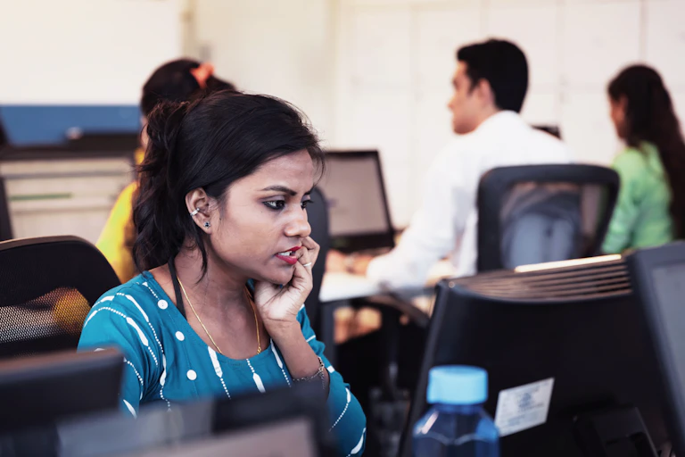 A woman looking worried at a busy office sitting at a row of computers.