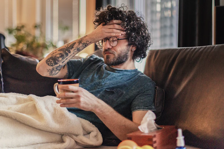 A man sitting on a couch under a blanket, with his hand on his forehead.