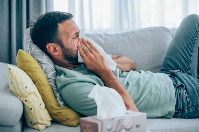 Man lies on couch blowing his nose.