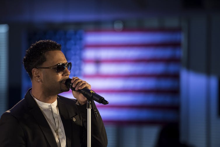 Man wearing suit and sunglasses sings into microphone with American flag hanging in background