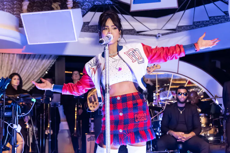 Woman in red and blue plais miniskirt, crop top and leather jacket sings into microphone