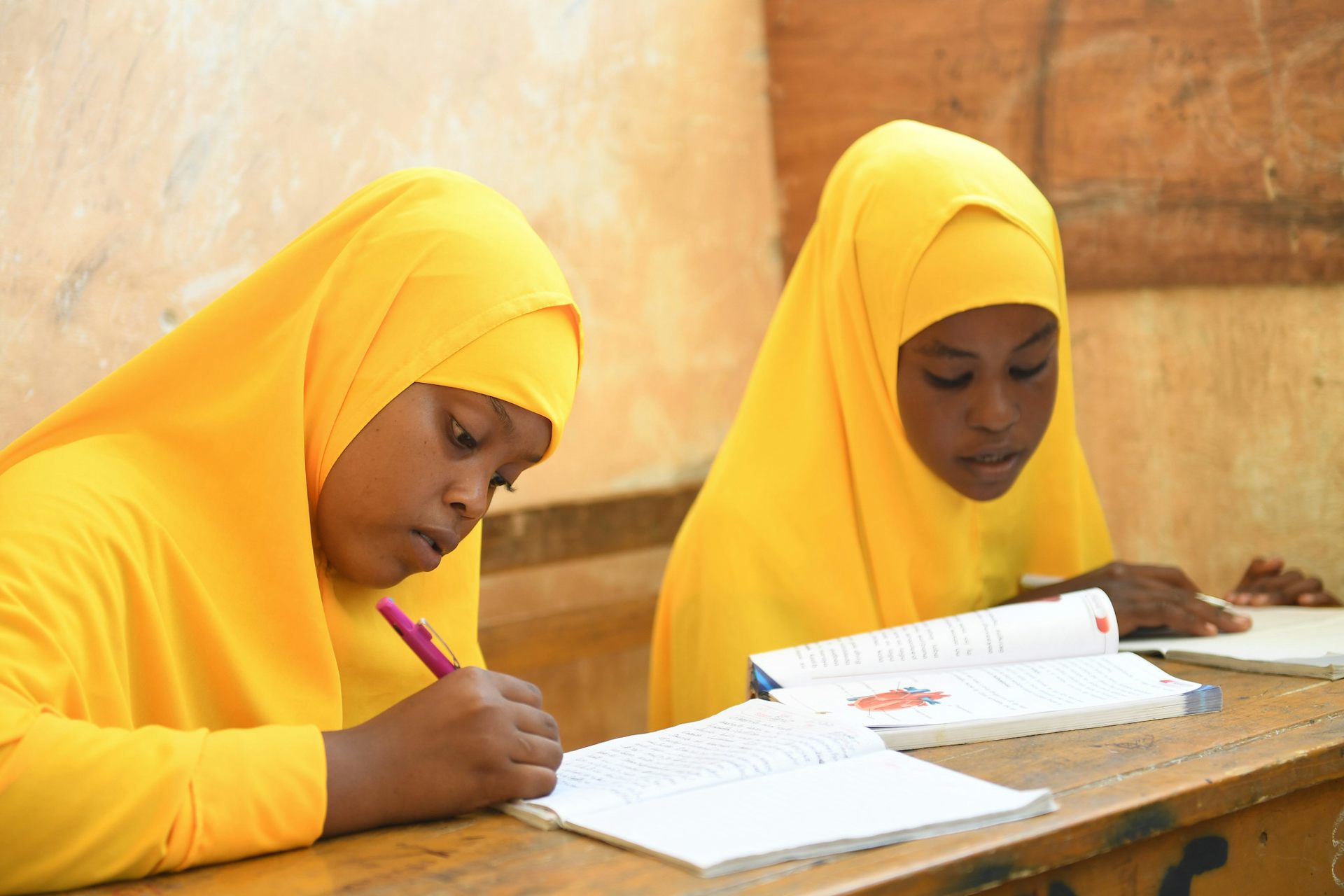 Two girls in yellow headscarves and clothing sit at a wooden desk with books