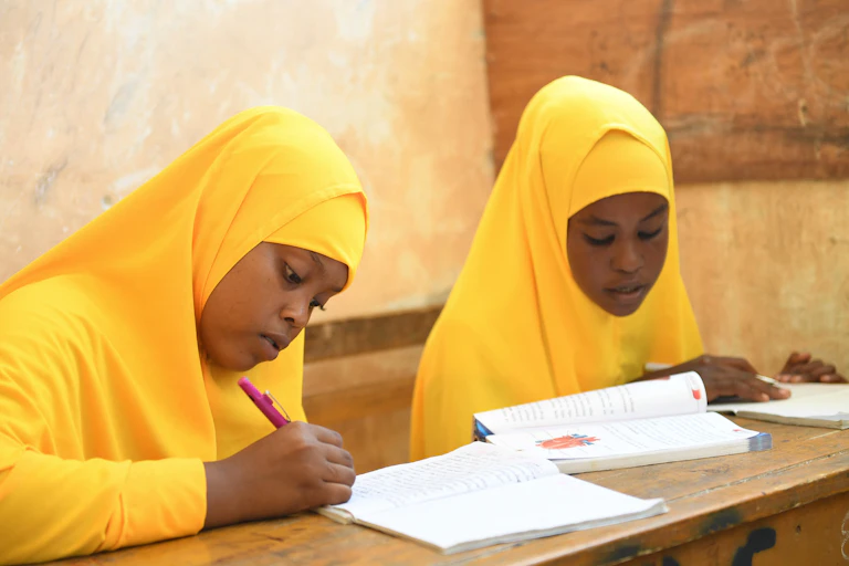 Two girls in yellow headscarves and clothing sit at a wooden desk with books