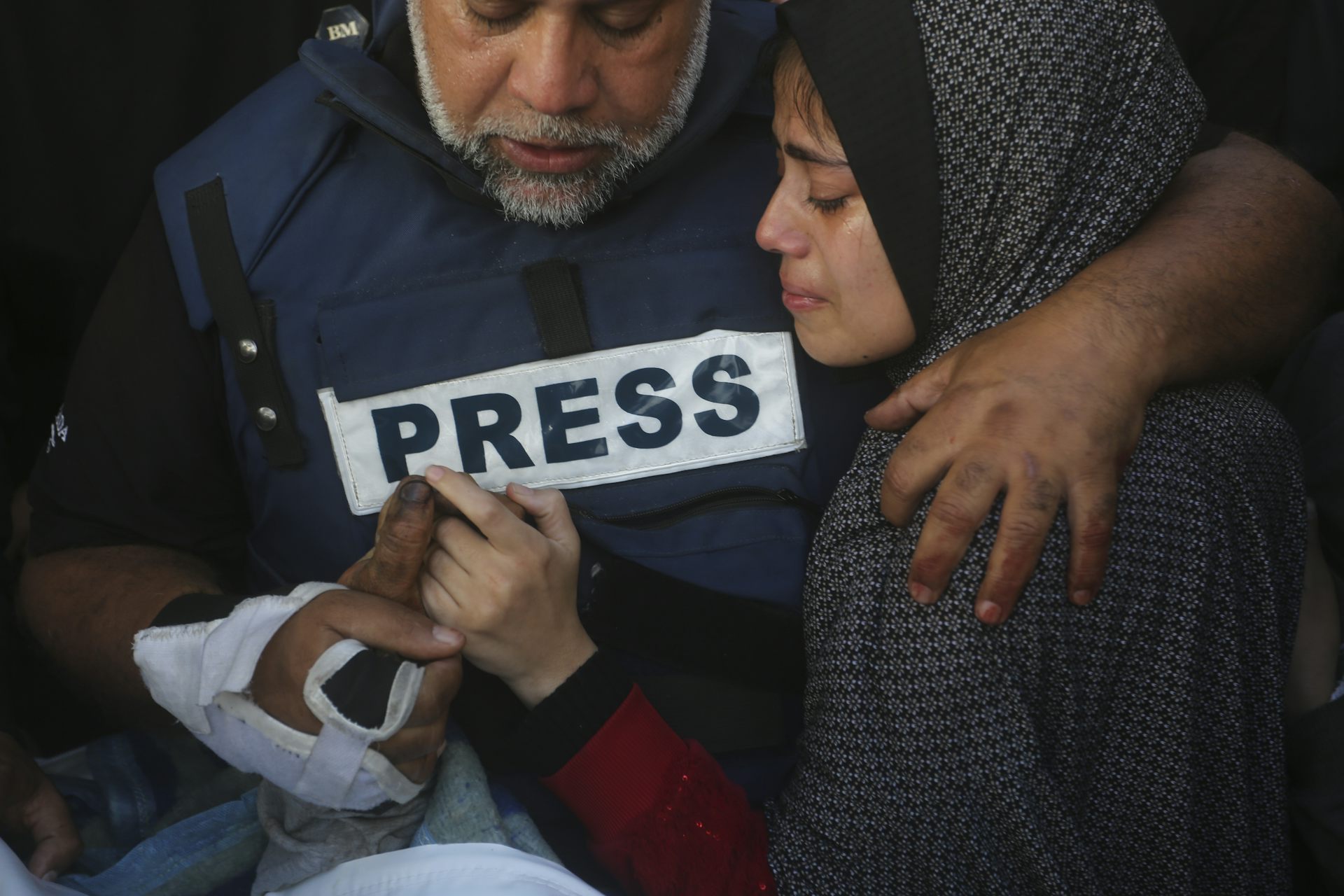A man holds the hand of his deceased son. A large sticker that says "press" is taped to his shirt.