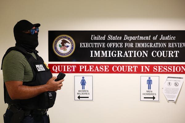 A man in a black mask, hat and vest stands in a hallway next to a sign that says 'IMMIGRATION COURT.'