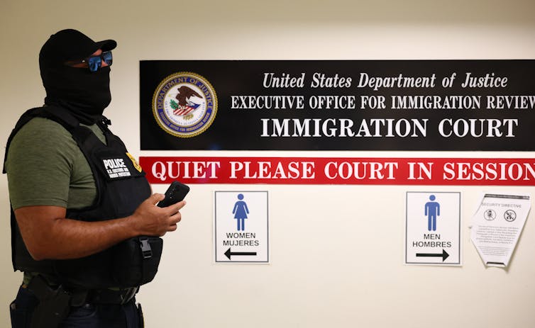 Një burrë me maskë të zezë, këpucë dhe xhaketë qëndron në një korridor pranë një shenje që thotë 'GJYKATA E MIGRACIONIT.'