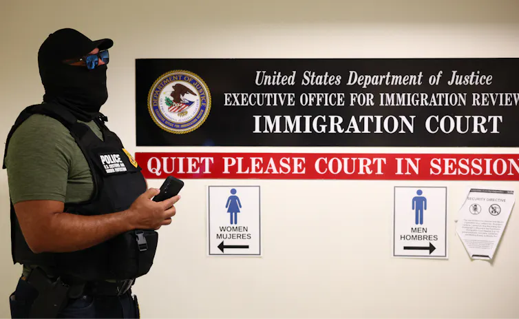 A man in a black mask, hat and vest stands in a hallway next to a sign that says 'IMMIGRATION COURT.'