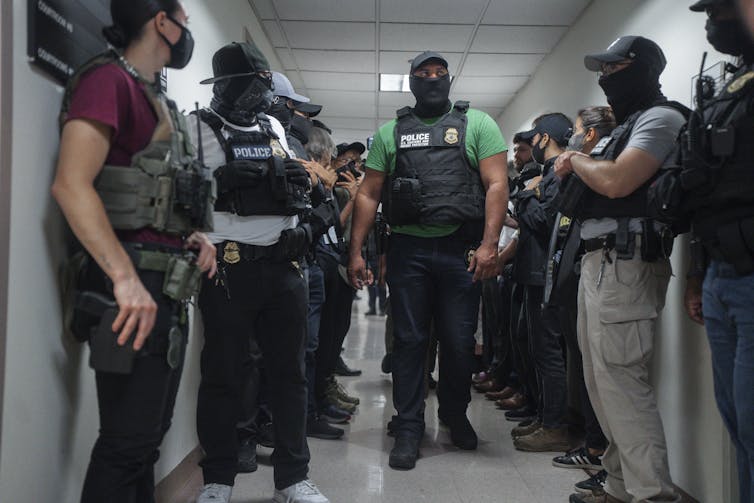 Masked men wearing hats and bulletproof vests, standing in a hallway.