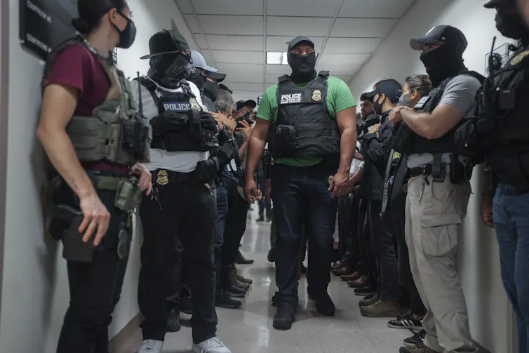 Masked men wearing hats and bulletproof vests, standing in a hallway.
