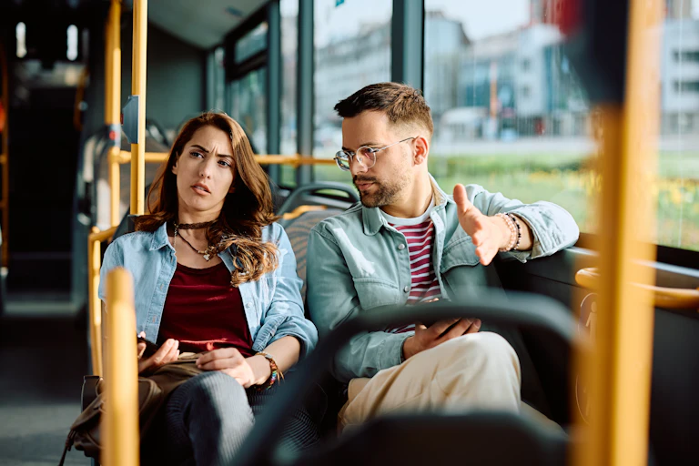 Un jeune couple hétérosexuel assis côte à côte dans un bus, la femme regarde ailleurs tandis que l'homme lui parle d'un ton sévère.