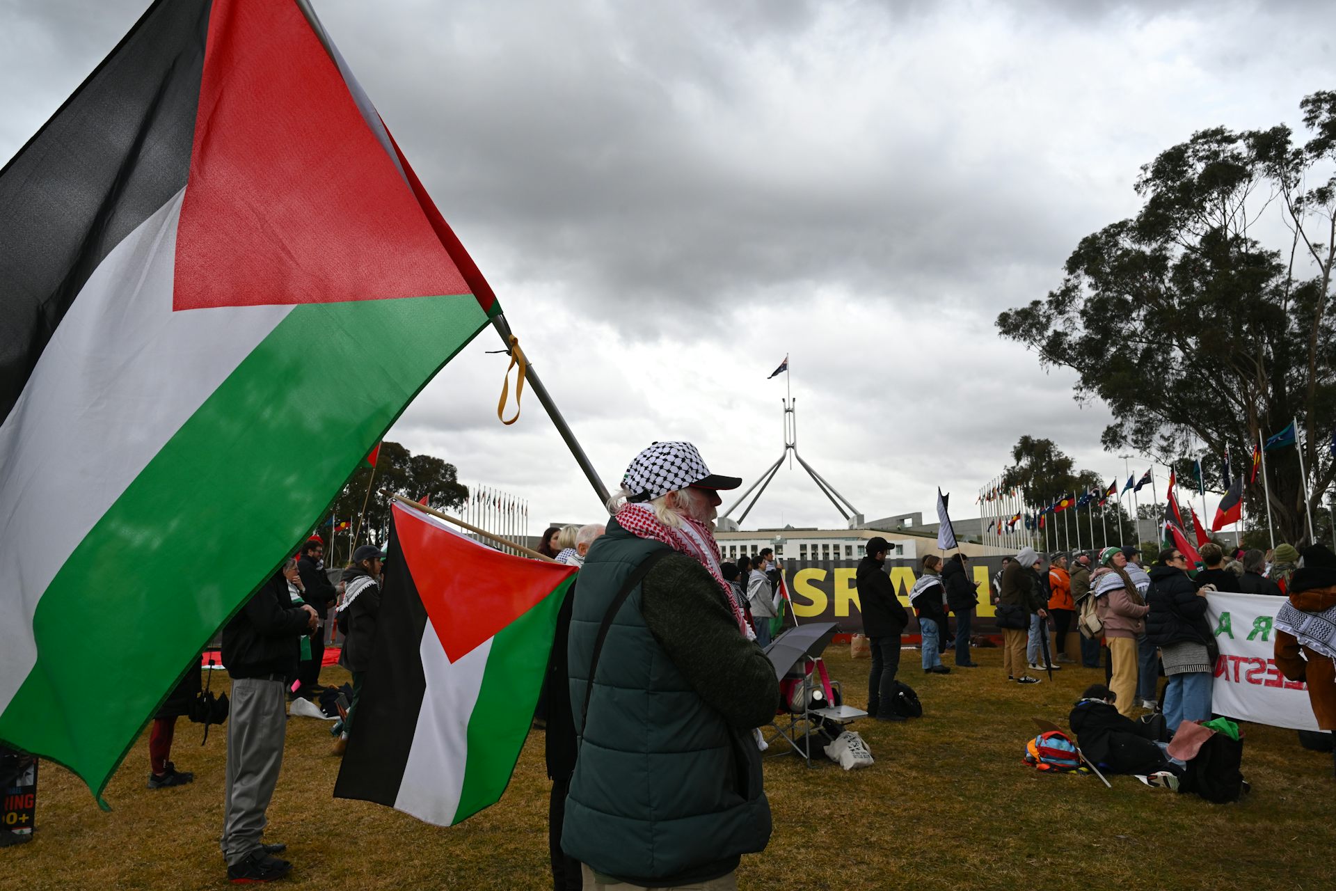 A man carrying a large Palestinian flag among protestors outside Parliament House in Canberra.