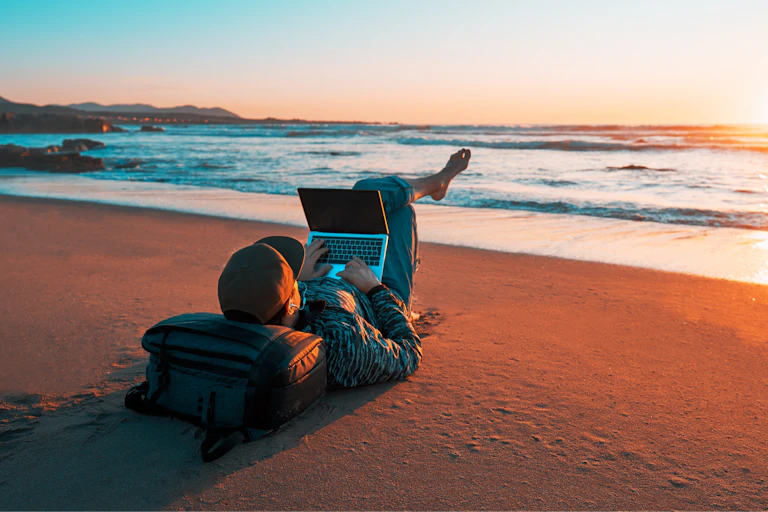 Traveller with laptop open on beach at sunset.