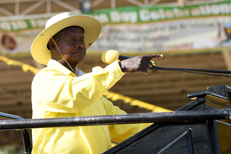 A man in a yellow shirt and wide-brim hat speaks into a microphone while pointing