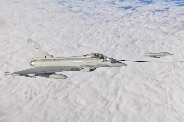 A military jet flies above the clouds with a refueling hose connected to it, stretching out of the picture.