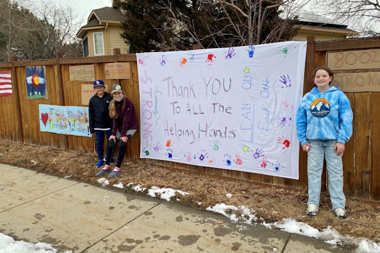 Children stand in front of a fence with a large sign that says "Thank you to all the helping hands."