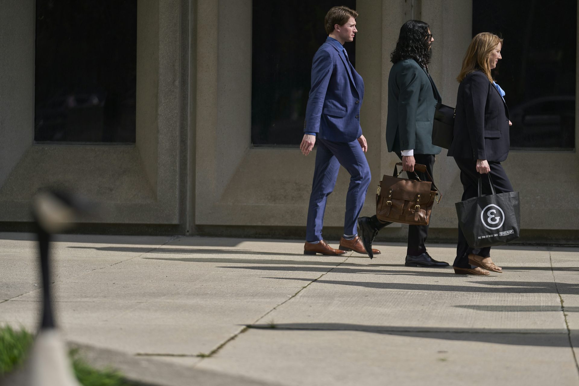 Un hombre con traje azul y dos mujeres caminan frente a la cancha fotografiadas en siluetas con sombras al lado de ellas.