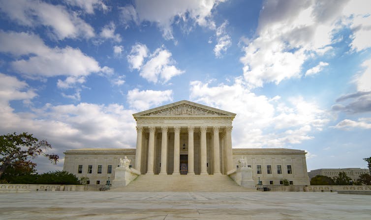 A large white, pillared building at the back of a plaza, with clouds in a blue sky behind it.