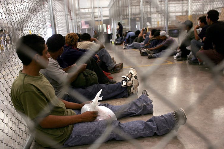 Several men seated on the ground are seen in a holding cell.
