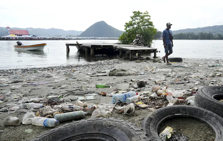 A man walks past plastic waste and discarded car tires strewn across a beach